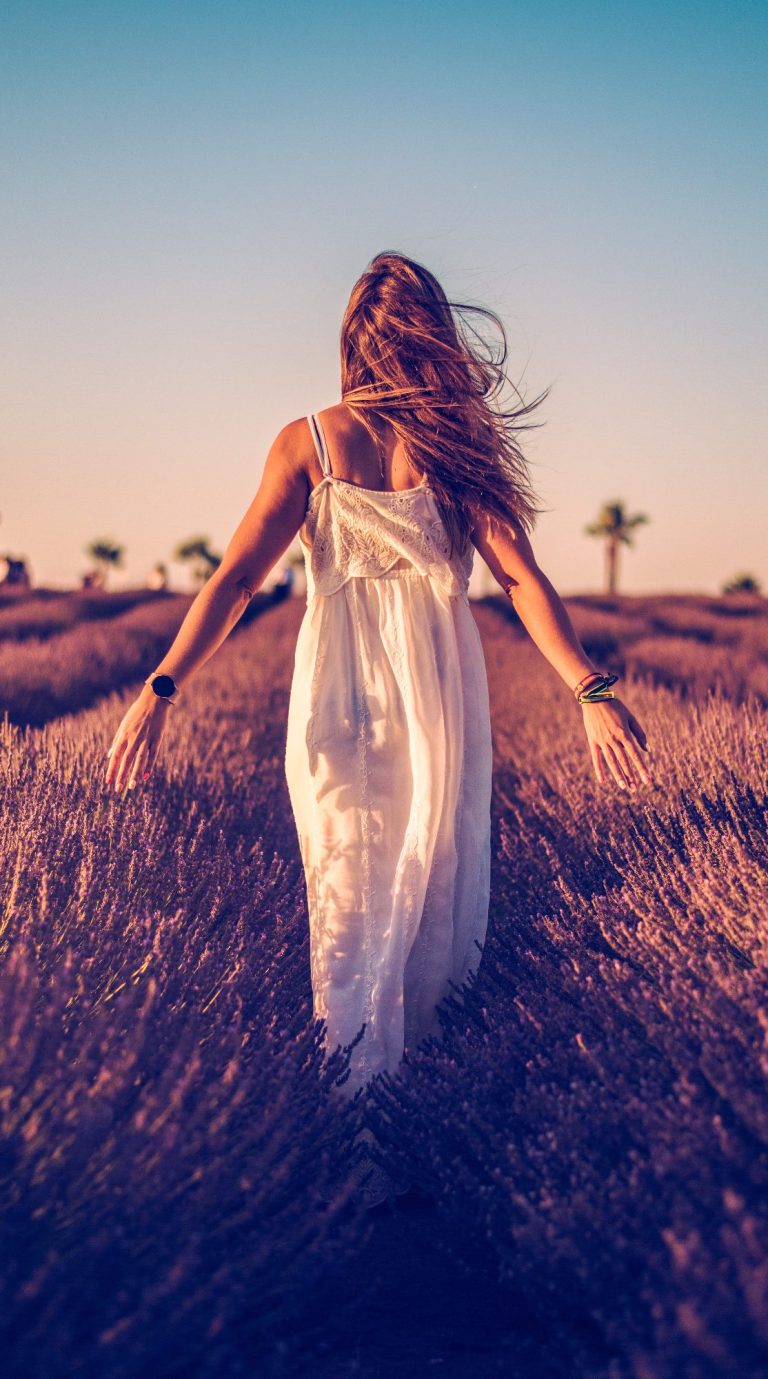 Mujer de espaldas caminando entre campos de flores al atardecer.