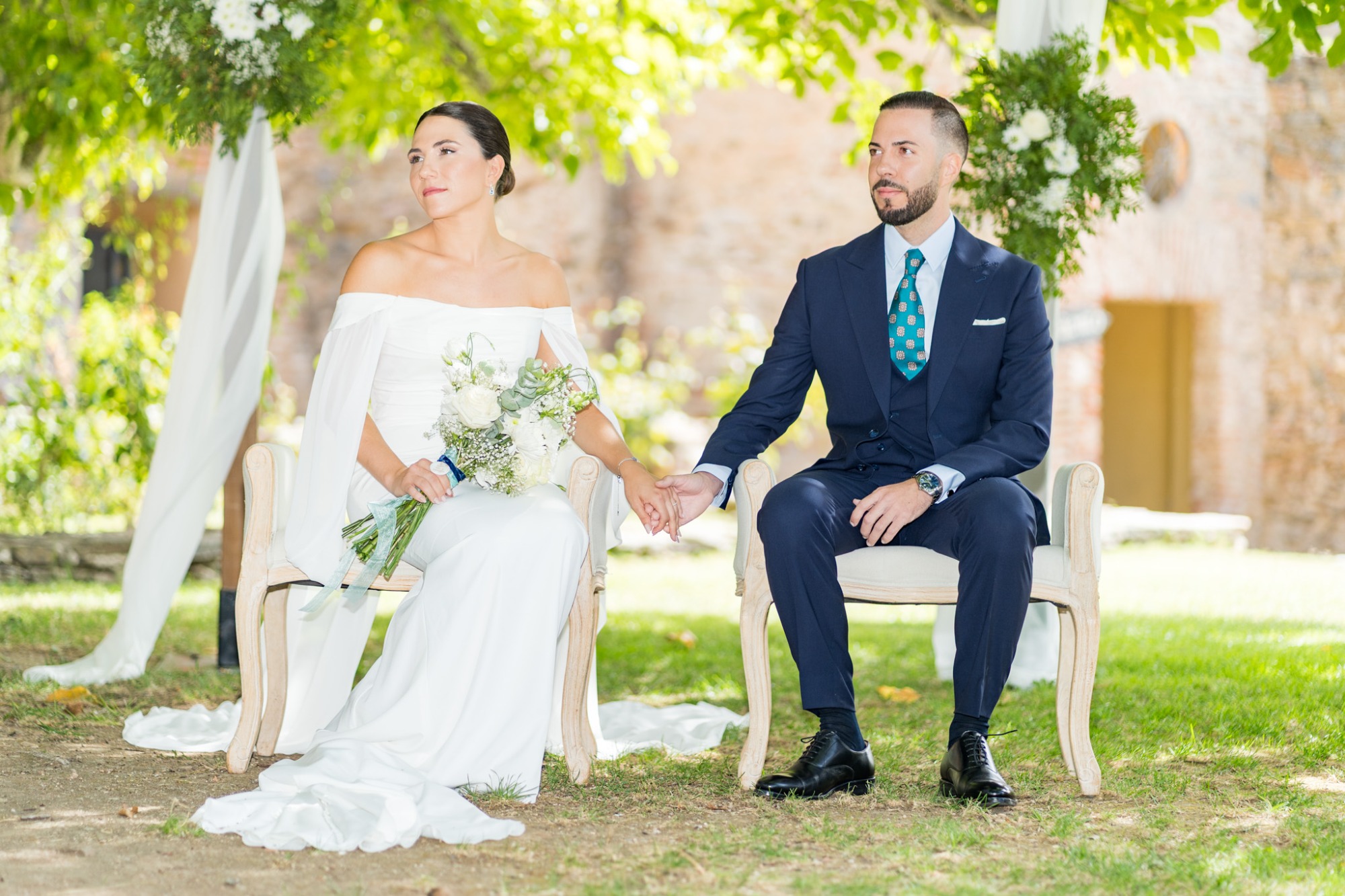 Pareja de novios sentados al aire libre, sosteniéndose de la mano bajo un árbol.