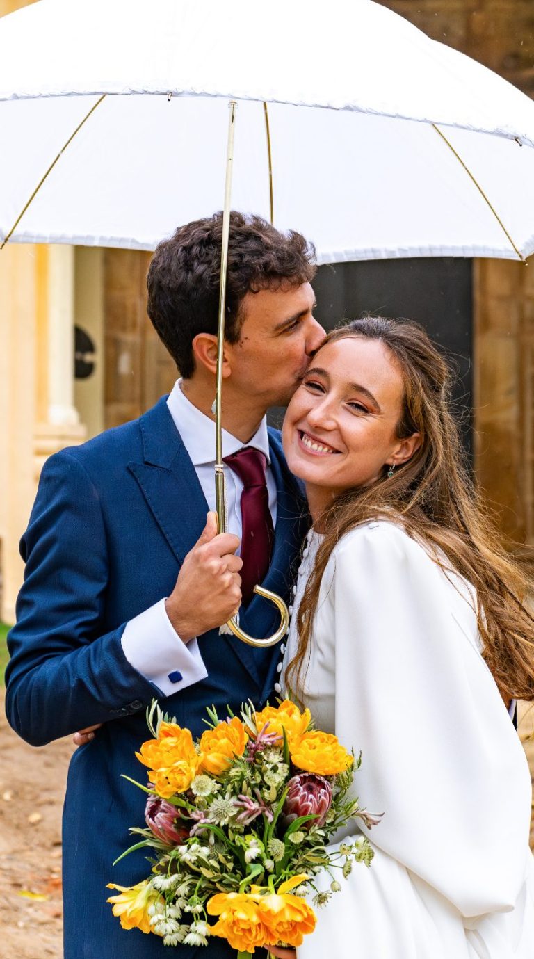 Pareja sonriente bajo un paraguas, con flores amarillas, en un entorno festivo.