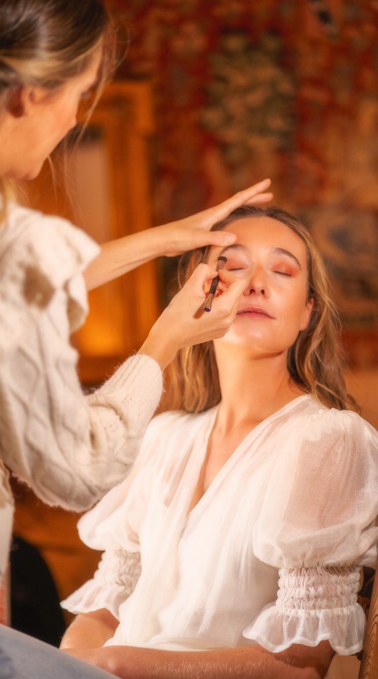 Una mujer recibe maquillaje de otra en un ambiente cálido y acogedor.