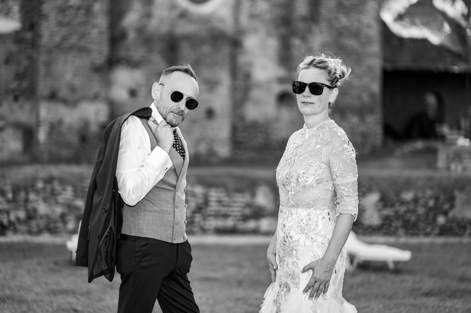 Pareja elegante en un entorno natural, luciendo gafas de sol y atuendos formales.