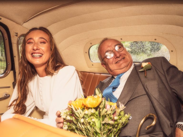 Joven sonriente con ramo de flores, sentada junto a un hombre mayor en un coche clásico.