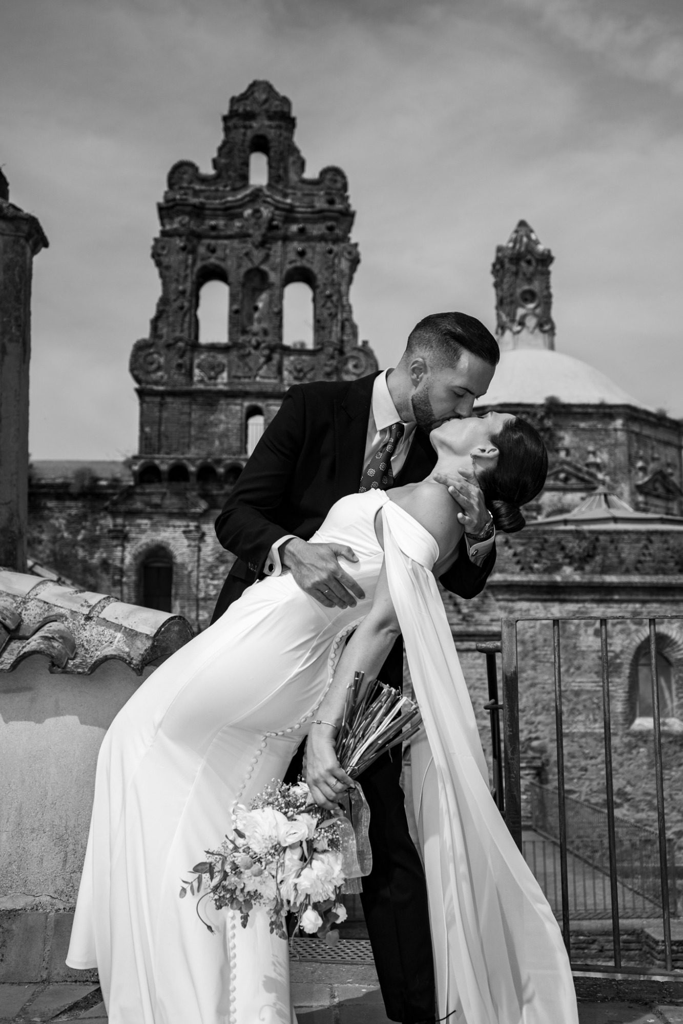Pareja de novios en una escena romántica, besándose con un fondo histórico y arquitectónico.