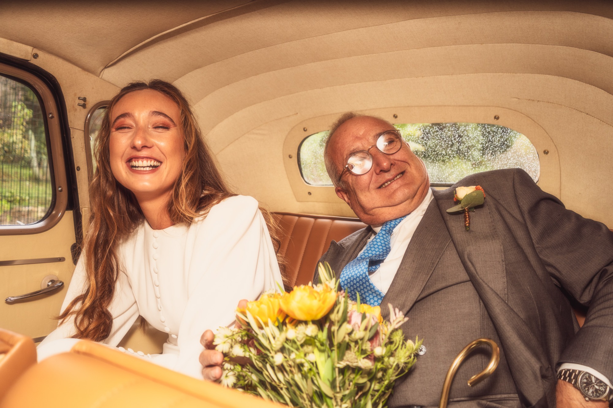 Mujer sonriendo con ramo de flores y hombre mayor en un coche clásico.
