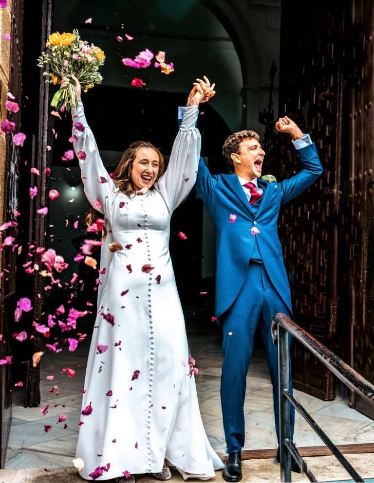 Pareja sonriente celebrando su boda, rodeada de pétalos y sosteniendo un ramo de flores.