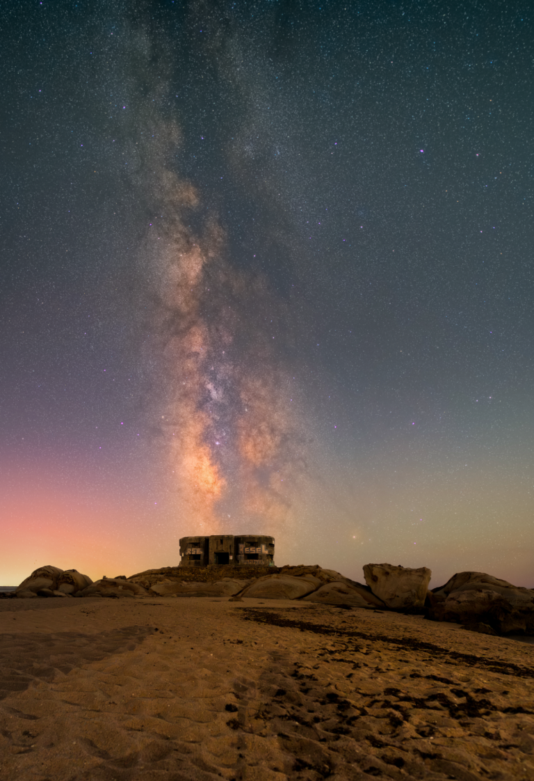 Cielo estrellado con la Vía Láctea sobre un paisaje rocoso y una construcción solitaria.