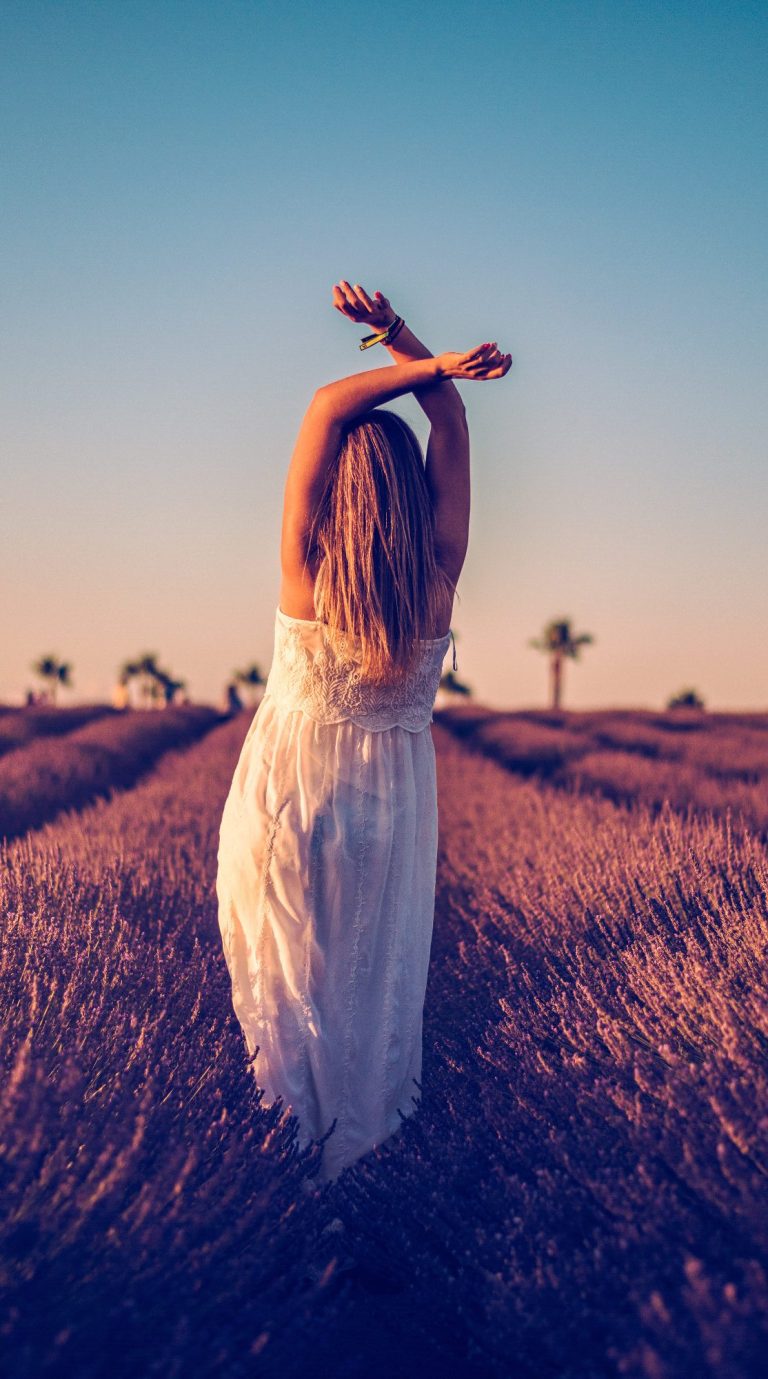 Mujer de espaldas en un campo de lavanda al atardecer, brazos en alto.