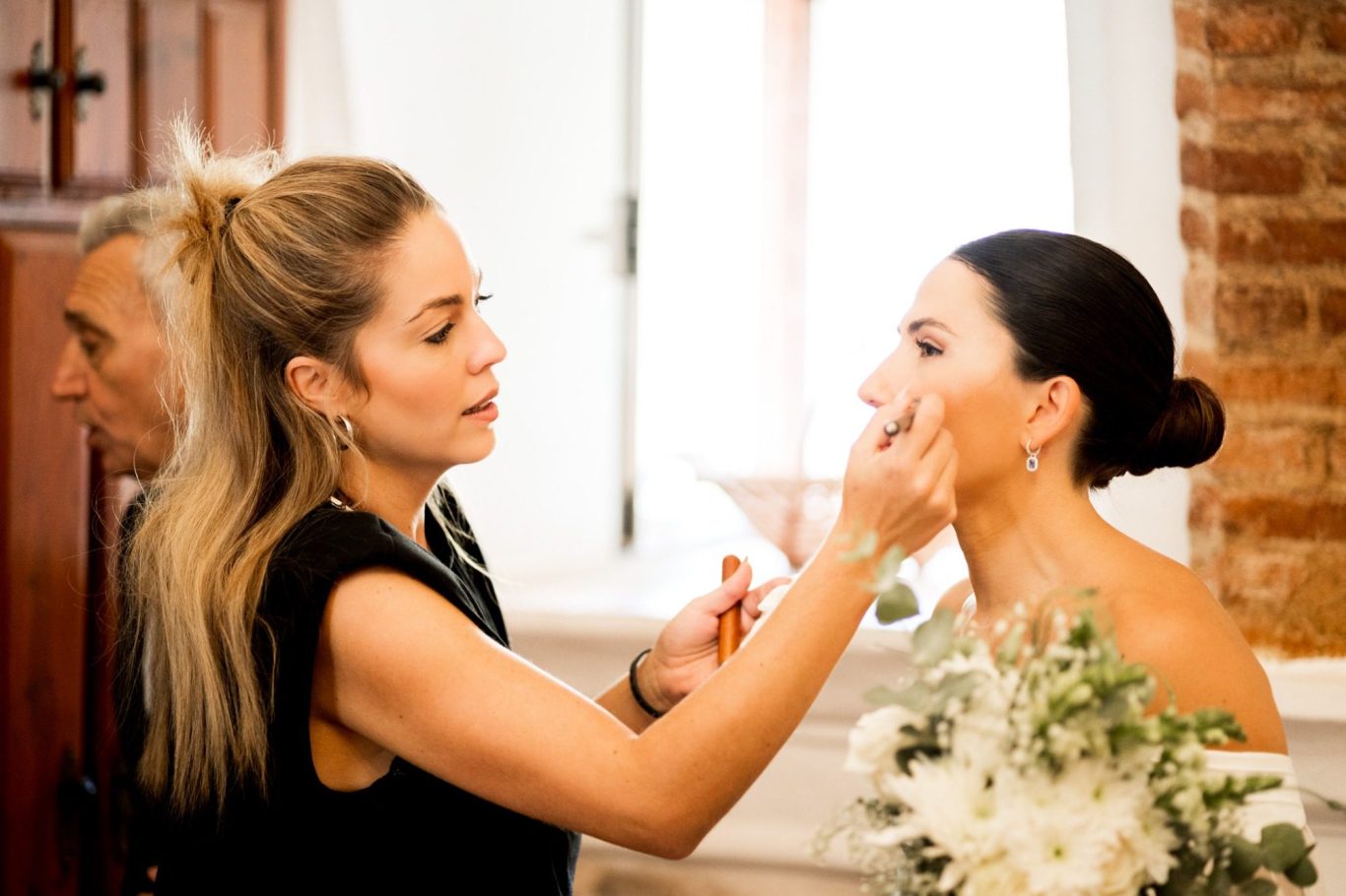 Mujer aplicando maquillaje a otra mujer, con bouquet de flores y fondo de ladrillo.