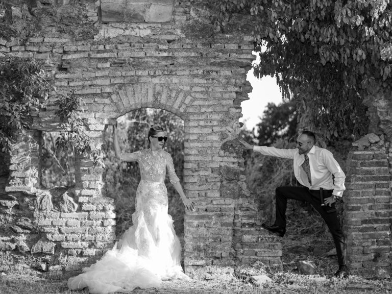 Novia en vestido blanco junto a un hombre en traje, frente a una pared de ladrillos.