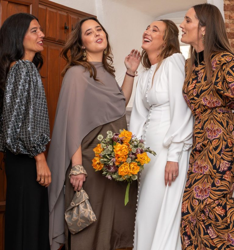 Cuatro mujeres sonrientes con vestidos elegantes, una sostiene un ramo de flores.