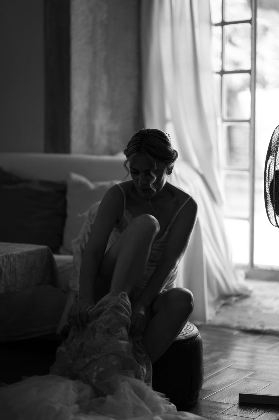 Novia sentada en un sofá poniéndose su vestido de novia, abrigada, con luz suave entrando por una ventana.