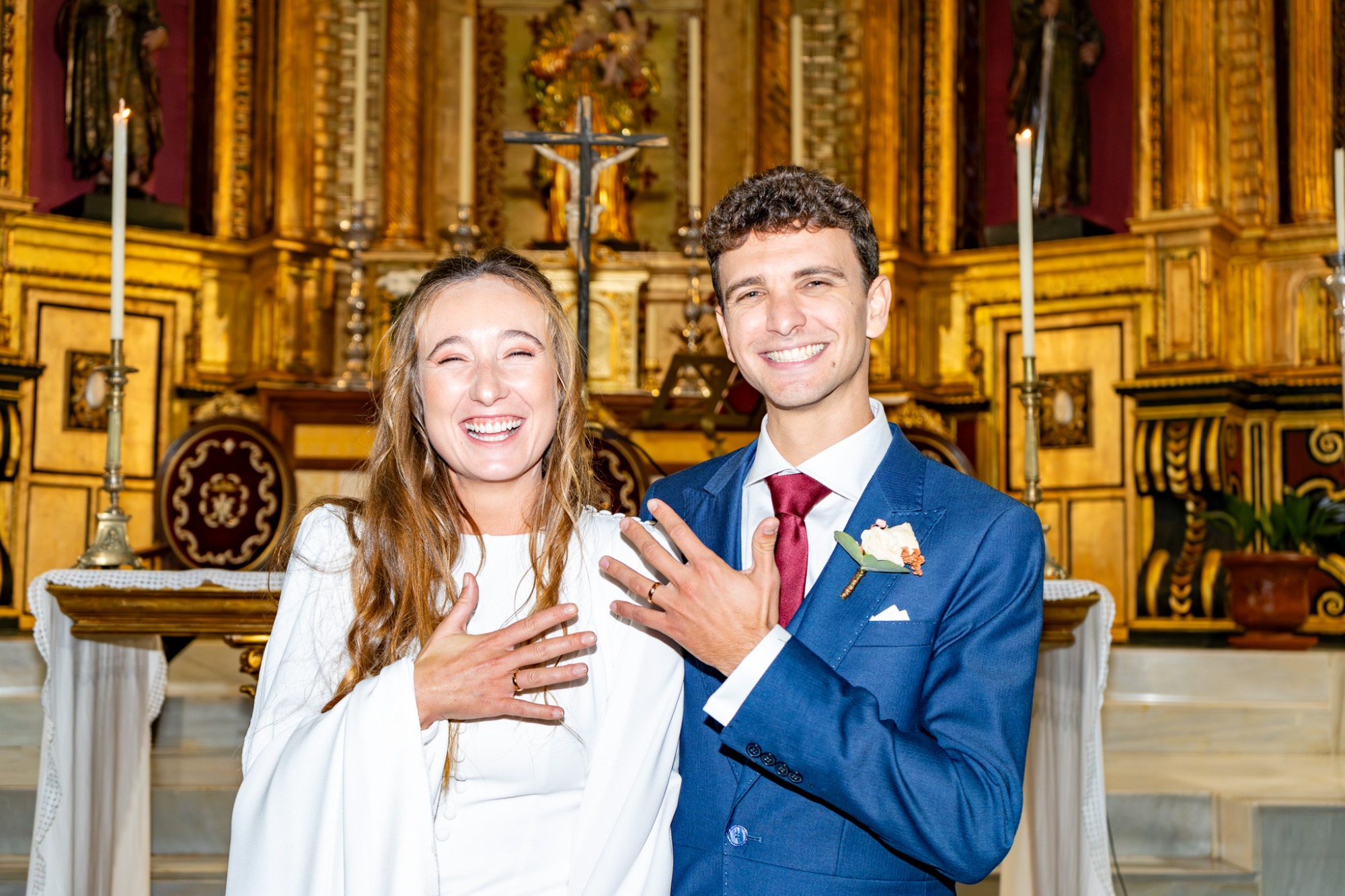 Pareja joven sonriente en un altar, vestida elegantemente, con decoraciones religiosas de fondo.